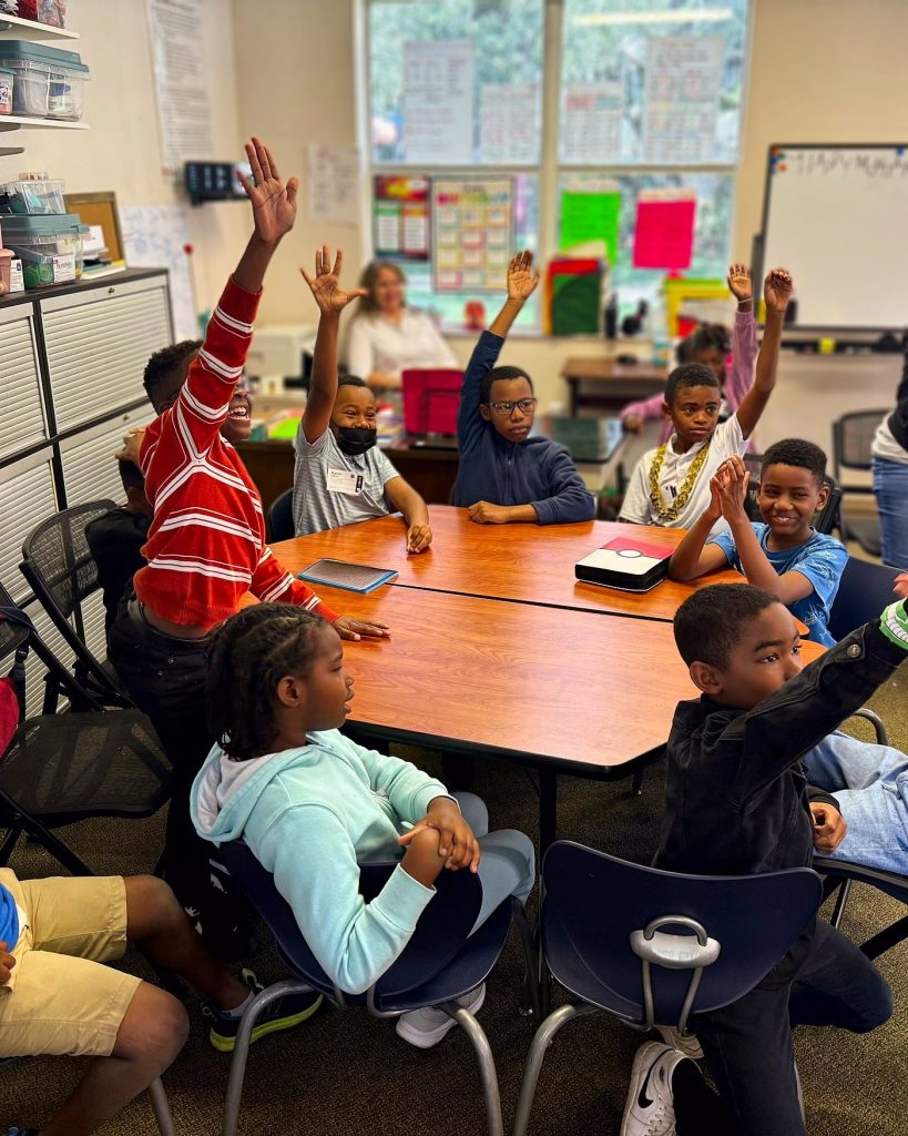 Kids raising their hands around a table in a One Family Kids classroom