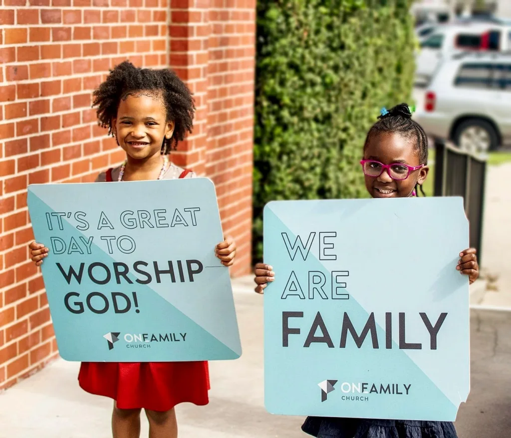 Two girls holding signs that say It's a great day to worship God and We are family