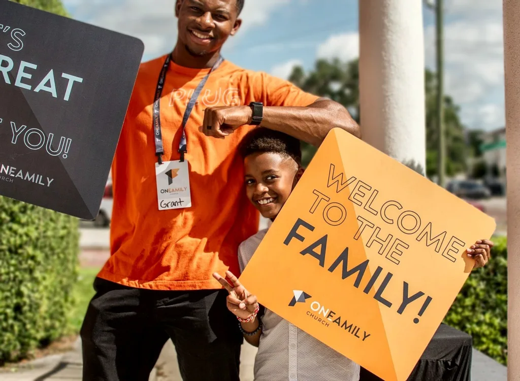 A greeter holding a Welcome to the Family sign with a smiling boy outside the church